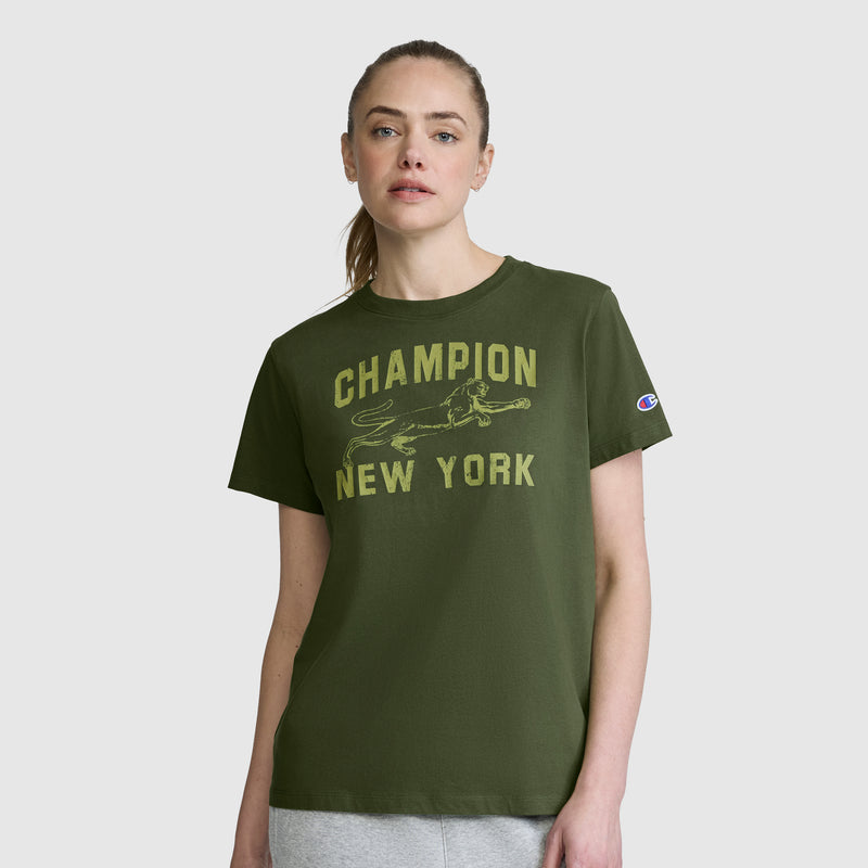 A woman stands against a plain light gray background wearing the Champion Mascot Graphic T-Shirt—an olive green women's tee featuring "CHAMPION NEW YORK" and a bold running dog graphic on the front.