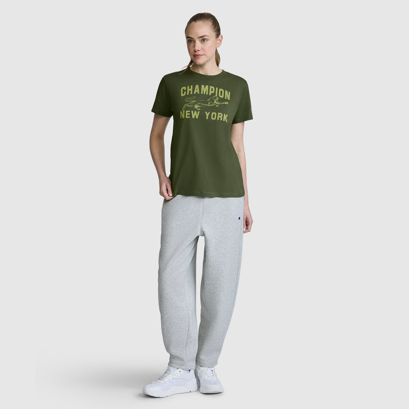 A woman stands against a plain background, wearing the Champion Mascot Graphic T-Shirt in green, paired with light gray sweatpants and white sneakers—a stylish women’s graphic tee from Champion.