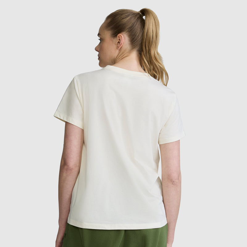 A woman with blonde hair in a ponytail, wearing the Champion Squad Graphic T-Shirt—crafted from lightweight cotton jersey—and green pants stands with her back to the camera against a plain light background.