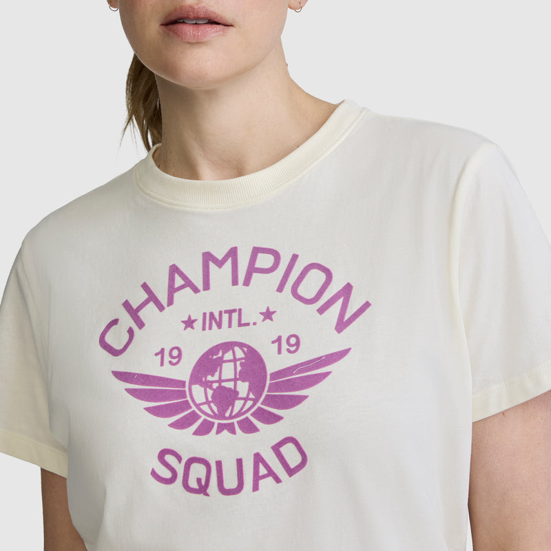 A person is shown from the lower face to upper torso, wearing the Champion Squad Graphic T-Shirt. This lightweight cotton jersey tee features bold purple text reading "CHAMPION SQUAD INTL. 1919" along with a globe with wings graphic, highlighting the collegiate style of this signature Champion piece.