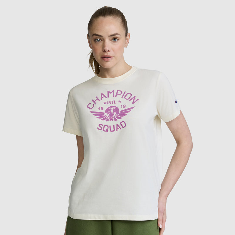 A woman stands against a plain background with one hand in her pocket, wearing the Champion Squad Graphic T-Shirt—a white tee featuring a collegiate-inspired purple winged basketball graphic. Made from lightweight cotton jersey, this shirt delivers all-day comfort and effortless style.