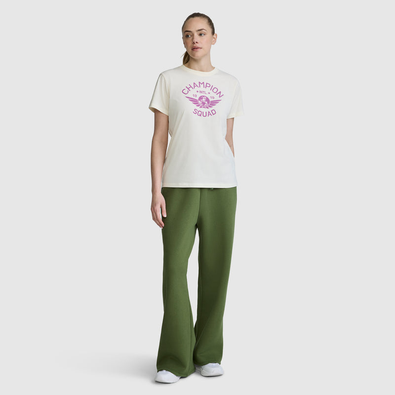 A woman stands against a plain white background wearing the Champion Squad Graphic T-Shirt, a lightweight cotton jersey tee with a white collegiate graphic and pink accents. She pairs it with wide-leg green pants and white sneakers.