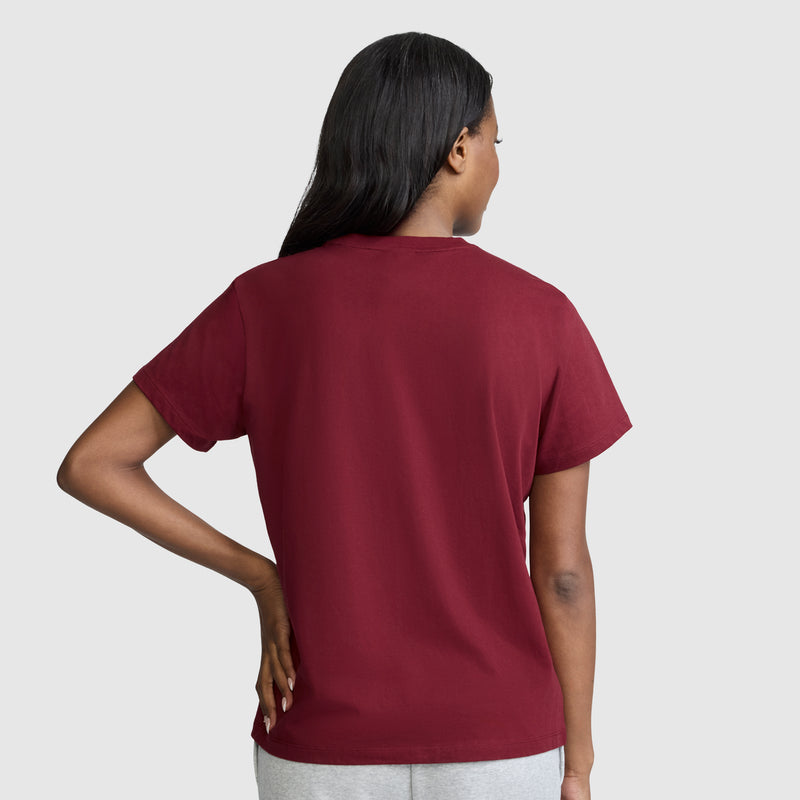 A person stands facing away, wearing the Champion New York Flower Graphic T-Shirt—a vintage collegiate-style, short-sleeve maroon tee—and light gray pants, with one hand resting on their hip. The background is light gray.