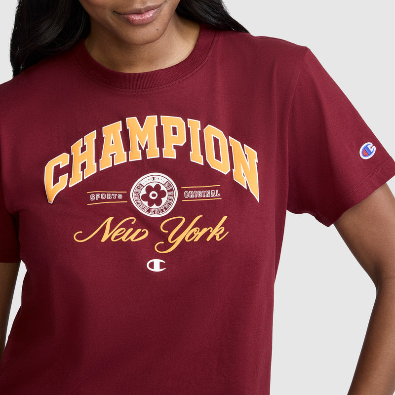 A person wearing a maroon Champion New York Flower Graphic T-Shirt, featuring vintage collegiate-style yellow and white text on the front. Only the upper body is visible.