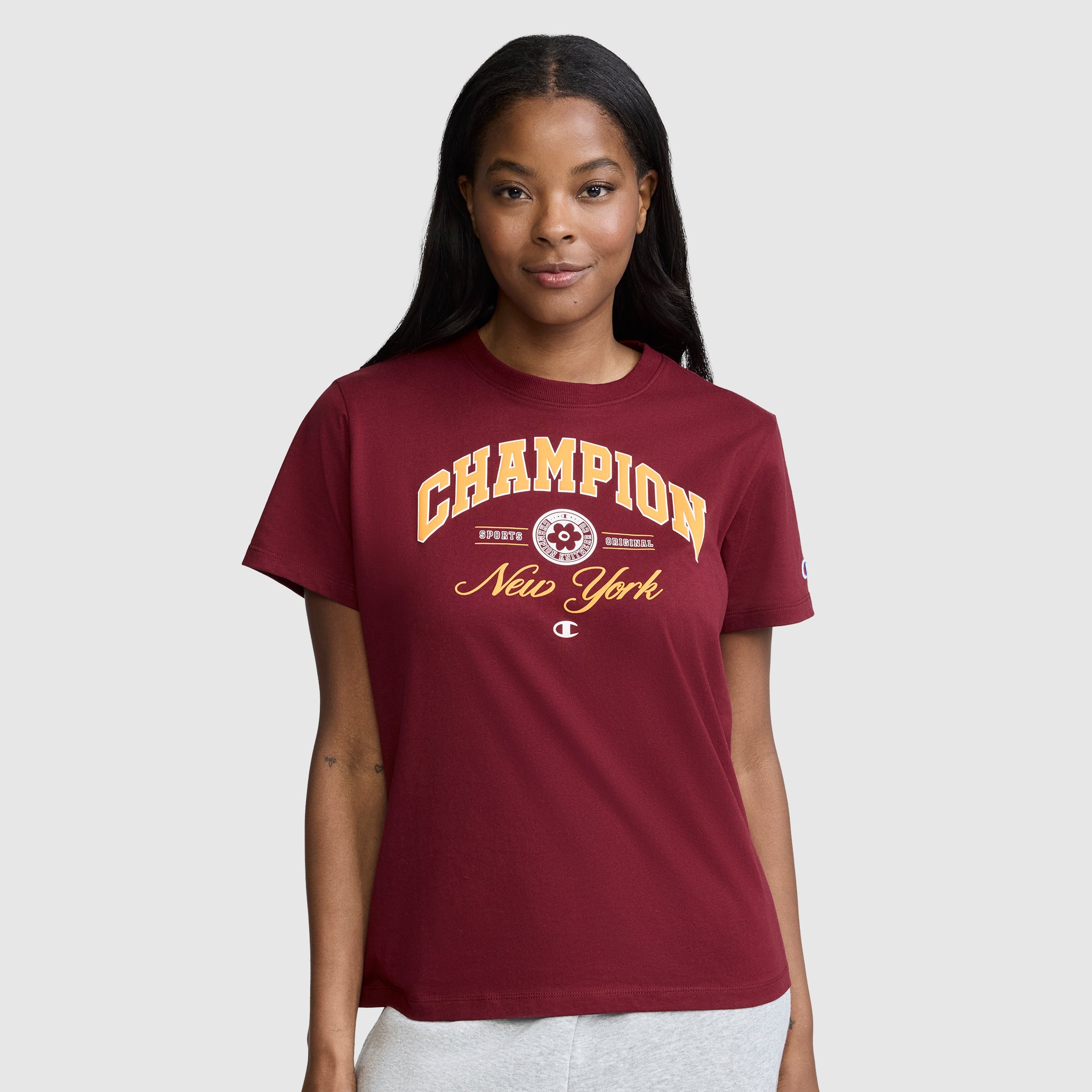 A woman wearing the Champion New York Flower Graphic T-Shirt in maroon, paired with light-colored pants, stands against a plain gray background and smiles softly at the camera.