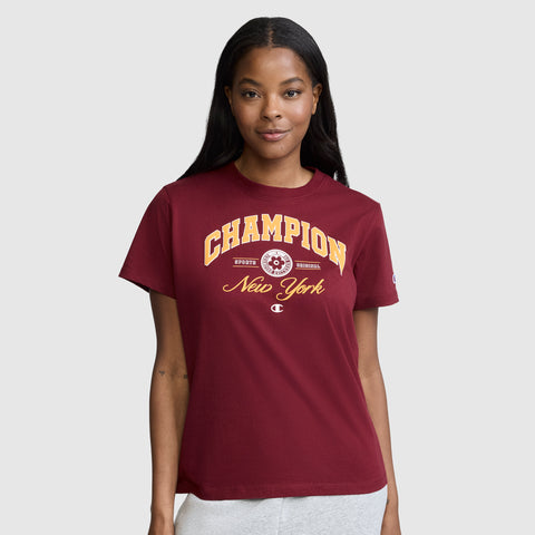 A woman wearing the Champion New York Flower Graphic T-Shirt in maroon, paired with light-colored pants, stands against a plain gray background and smiles softly at the camera.