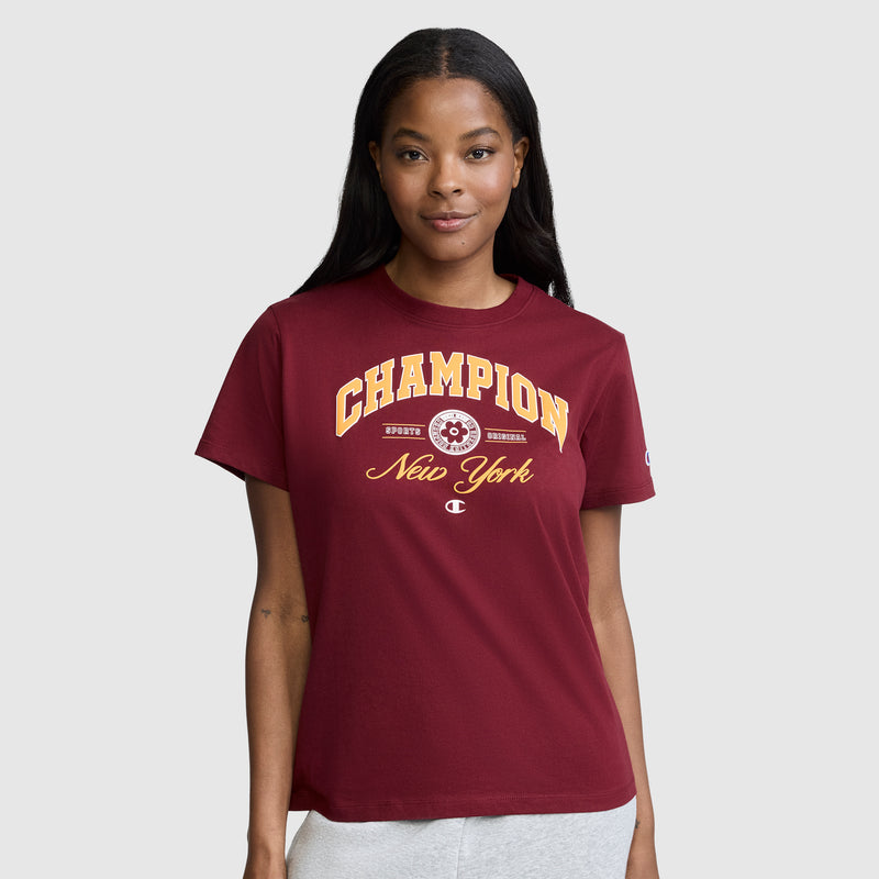A woman wearing the Champion New York Flower Graphic T-Shirt in maroon, paired with light-colored pants, stands against a plain gray background and smiles softly at the camera.