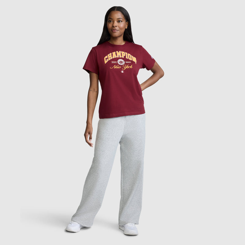 A woman stands and smiles, wearing a maroon Champion New York Flower Graphic T-Shirt with a vintage collegiate style, paired with light gray sweatpants and white sneakers against a plain light gray background.