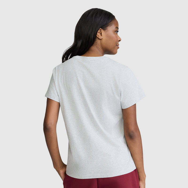 A woman with long dark hair is wearing a light gray Champion Sports Club "19" with Stars Graphic T-Shirt and red pants, standing with her back to the camera against a plain light background.