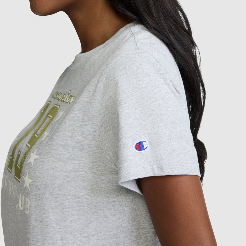 A woman wearing a light gray Champion Sports Club "19" with Stars Graphic T-Shirt featuring a gold graphic on the front. This vintage athletic tee showcases the Champion logo patch on the sleeve and is made from breathable cotton for all-day comfort. The background is plain light gray.