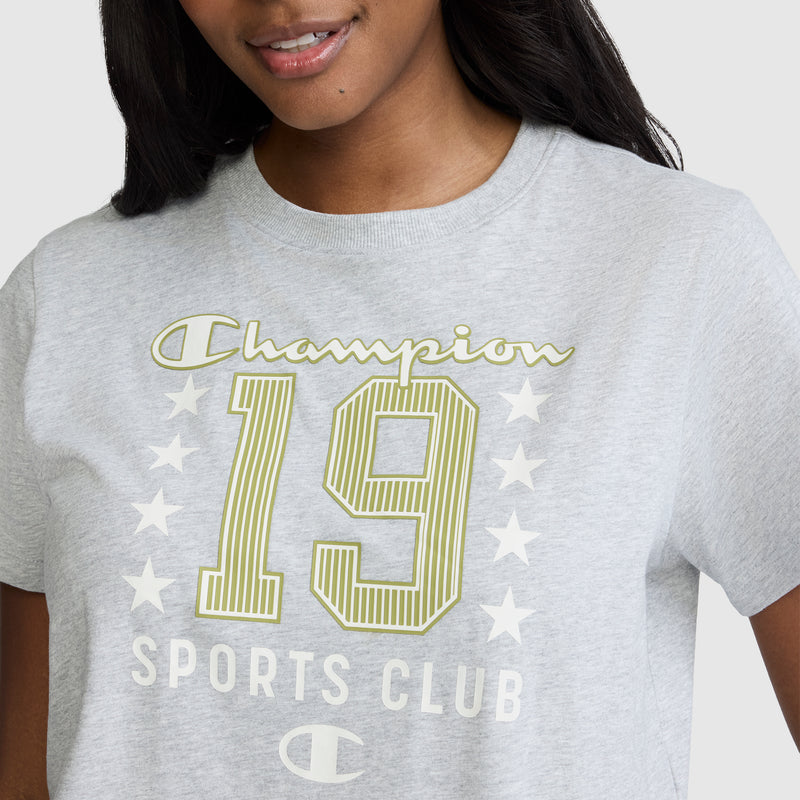 A woman wearing a light gray Champion Sports Club "19" with Stars Graphic T-Shirt. Featuring the words "Champion 19 Sports Club" and white stars printed on the front, this Champion tee blends vintage athletic style and comfort—perfect for casual days.