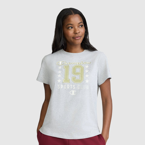 A woman is wearing the Champion Sports Club "19" with Stars Graphic T-Shirt in light gray, featuring a front print of "Champion 19 Sports Club" with stars. She stands with her hands in her pockets, looking slightly to the side. This vintage-style t-shirt by Champion is crafted from breathable cotton fabric for all-day comfort.