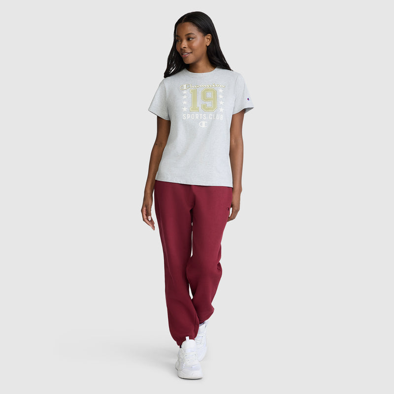 A woman wearing the Champion Sports Club "19" with Stars Graphic T-Shirt in light gray, paired with burgundy sweatpants and white sneakers, walks confidently against a plain white background.