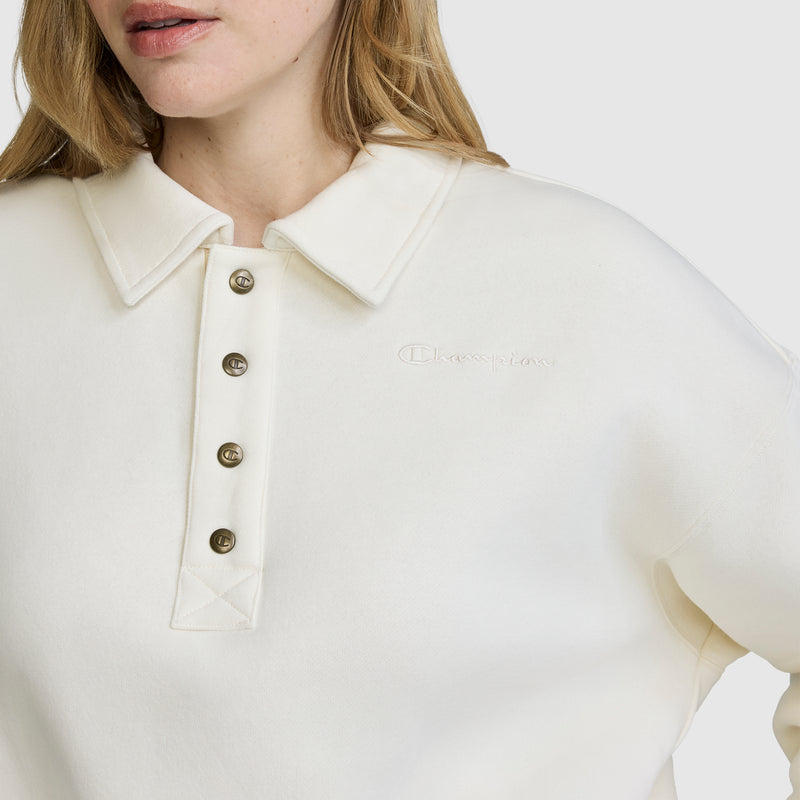 A woman is seen wearing the Champion Fleece Training Polo in off-white, featuring four buttons and an embroidered Champion logo on the chest. The photo focuses on her shoulders and lower face, emphasizing the stylish comfort of this women's polo shirt.