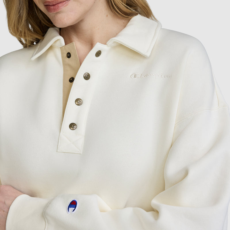 A woman with light hair is seen against a plain background, modeling the Champion Fleece Training Polo. This cream-colored pullover features metal buttons, an embroidered Champion logo on the chest, and a signature patch on the sleeve, highlighting its stylish athletic design.
