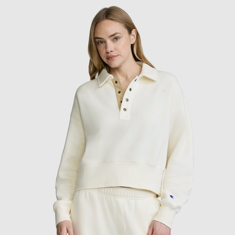 A woman wearing the Champion Fleece Training Polo, an off-white athletic pullover with a collared neckline and buttons, paired with matching pants, stands against a plain light background, looking at the camera with a neutral expression.