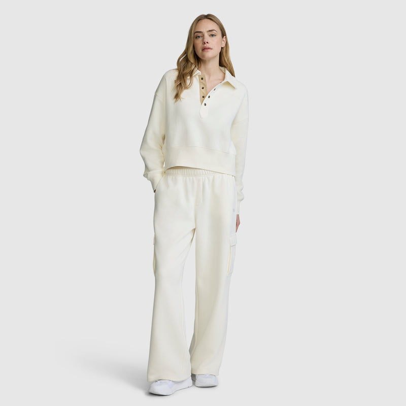 A person is wearing a cream-colored Champion Fleece Training Polo featuring long sleeves and buttons, styled with matching wide-leg pants. They have long, light brown hair and white sneakers, standing against a plain background.