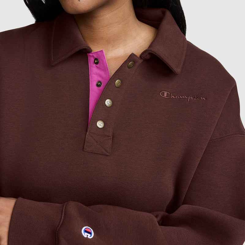 A person is wearing the Champion Fleece Training Polo in brown, featuring a collar, pink inner placket, and metallic buttons. Only the torso along with part of the face and arm are visible, highlighting this stylish women’s pullover that offers an athletic yet casual look.