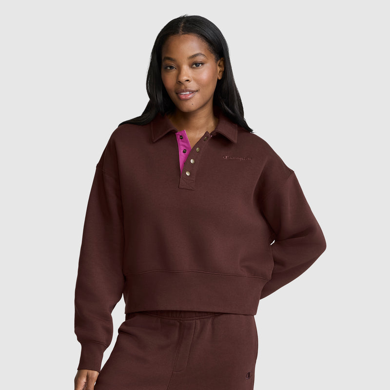 A woman stands against a light gray background, smiling with one hand on her hip, dressed in a Champion Fleece Training Polo featuring a magenta collar and matching pants.