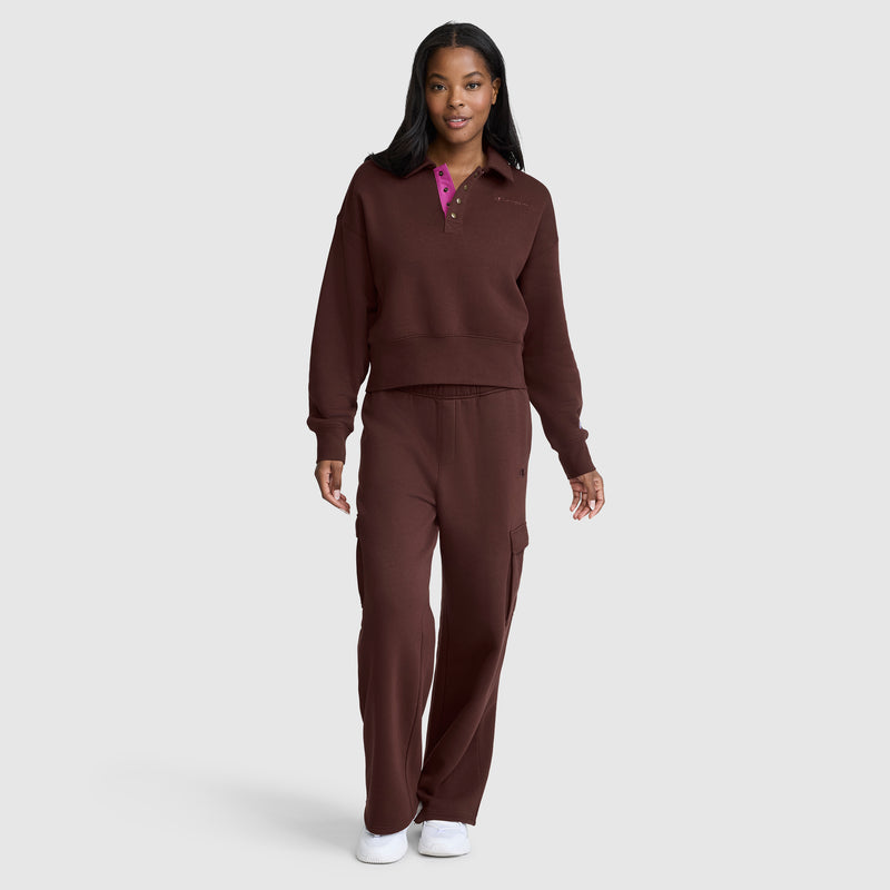 A woman stands against a plain light gray background, wearing the Champion Fleece Training Polo paired with matching pants and white sneakers.