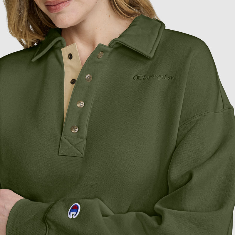 A woman wears a Champion Fleece Training Polo in olive green, featuring a beige collar and button placket, with the Champion logo displayed on the chest and sleeve. This women's pullover blends classic style and athletic polo details for a comfortable, sporty look.