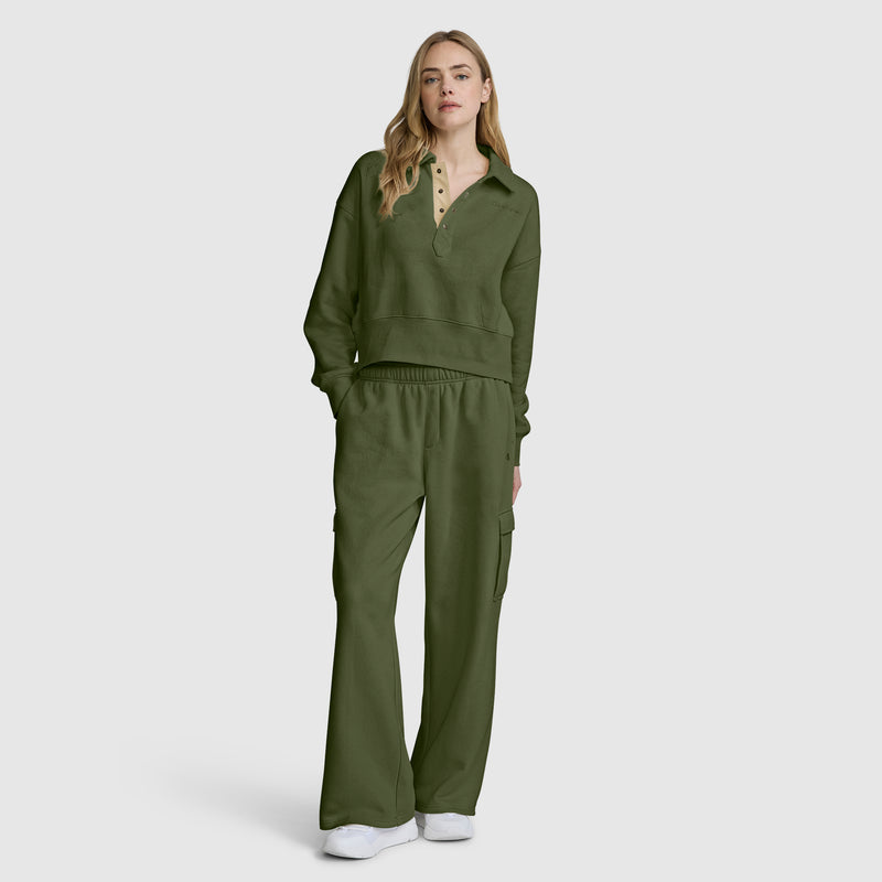 A woman stands facing forward, wearing a Champion Fleece Training Polo in olive green paired with matching wide-leg pants. She has long blonde hair and white sneakers, and is posing against a plain light gray background.