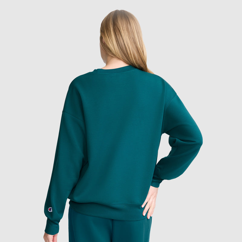 A person with long blonde hair, seen from behind, wears a teal Champion Crewneck Pullover Sweatshirt and matching pants, creating an athletic silhouette. Their right hand rests on their hip against a light gray background.