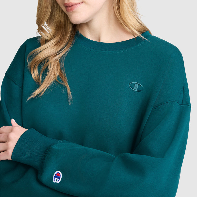 A woman wears a Champion Crewneck Pullover Sweatshirt in teal, showcasing an athletic silhouette and visible brand logos on the chest and sleeve, set against a plain light gray background.