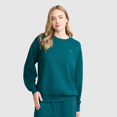 A woman with long blonde hair wears a teal Crewneck Pullover Sweatshirt and matching pants by Champion, standing against a light gray background and looking at the camera with a neutral expression. The athletic silhouette of the Champion Crewneck Pullover Sweatshirt adds a modern touch to her relaxed ensemble.