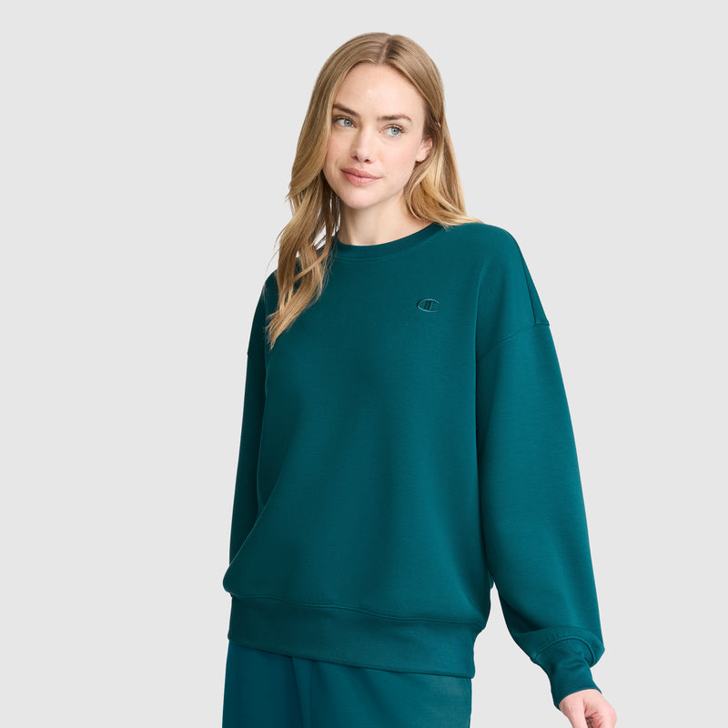 A woman with long blonde hair wears a teal Champion Crewneck Pullover Sweatshirt and matching pants, standing against a plain light gray background while looking slightly to the side and smiling gently. The athletic silhouette of her outfit lends a modern touch to her relaxed appearance.