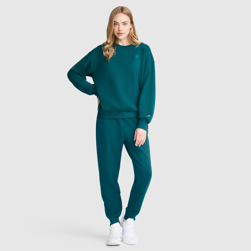 A woman with long blonde hair stands against a plain background, highlighting an athletic silhouette in a matching teal Champion Crewneck Pullover Sweatshirt and sweatpants set paired with white sneakers. One hand rests on her hip while the other arm hangs relaxed by her side.