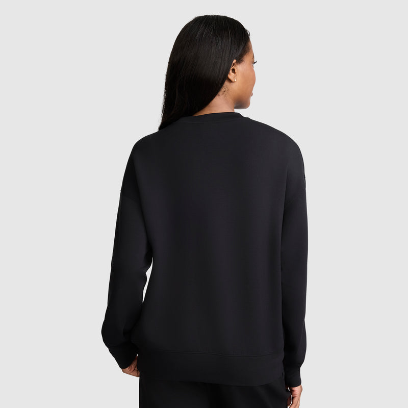 A woman with long dark hair stands with her back to the camera against a light gray background, wearing black pants and a Champion Crewneck Pullover Sweatshirt. The plain black modal scuba design of the sweatshirt creates an athletic silhouette.