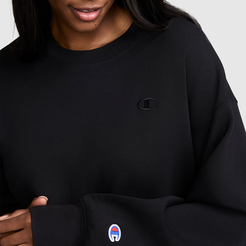 A person is shown from the shoulders to mid-torso wearing the Champion Crewneck Pullover Sweatshirt in black, featuring an embroidered C logo on the chest and a second logo on the sleeve, showcasing a classic athletic silhouette.