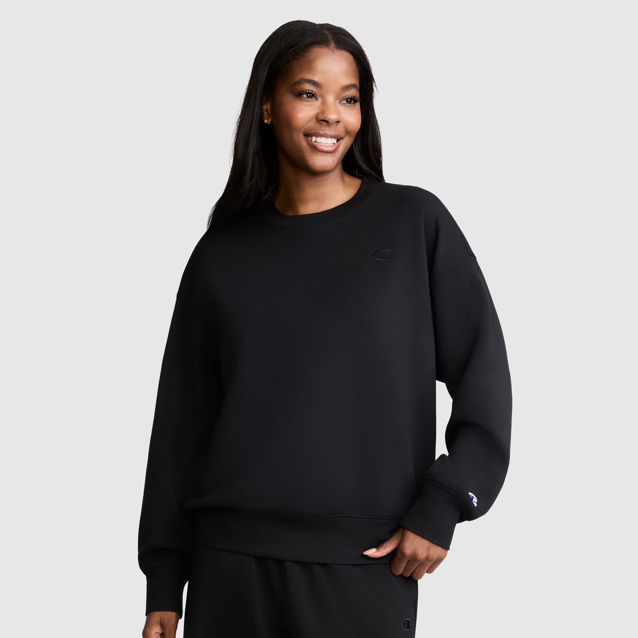 A woman with long dark hair wears a loose black Crewneck Pullover Sweatshirt by Champion, paired with matching pants. She stands against a plain light gray background and smiles slightly to the right. The relaxed fit of her Champion sweatshirt offers an effortlessly athletic silhouette.