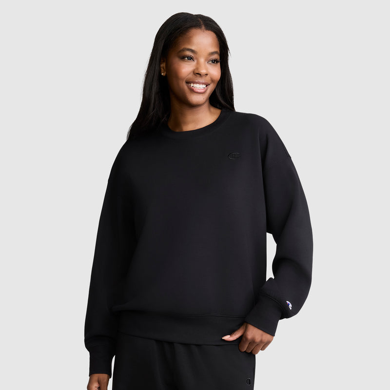 A woman with long dark hair wears a loose black Crewneck Pullover Sweatshirt by Champion, paired with matching pants. She stands against a plain light gray background and smiles slightly to the right. The relaxed fit of her Champion sweatshirt offers an effortlessly athletic silhouette.