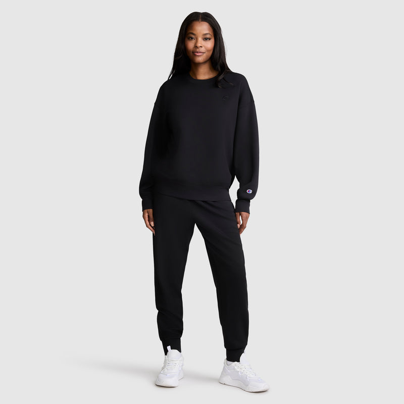 A woman stands facing forward, wearing a Champion Crewneck Pullover Sweatshirt in black with matching joggers and white sneakers, showcasing an athletic silhouette against a plain light gray background.