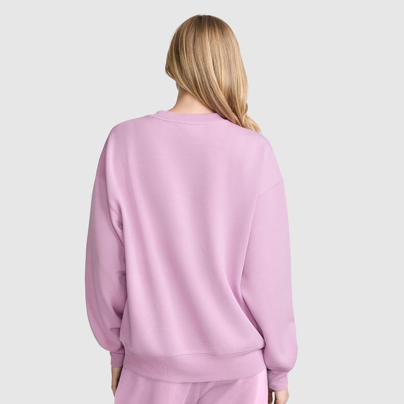 A person with long blonde hair is shown from the back, wearing a loose-fitting, athletic silhouette Champion Crewneck Pullover Sweatshirt and matching pants, standing against a plain light background.