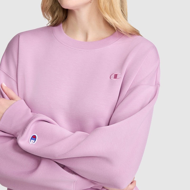 A blonde-haired woman wears the Champion Crewneck Pullover Sweatshirt, which features an athletic silhouette. Her arms are crossed, and the Champion logo is visible on both the chest and sleeve. The background is a plain light gray.
