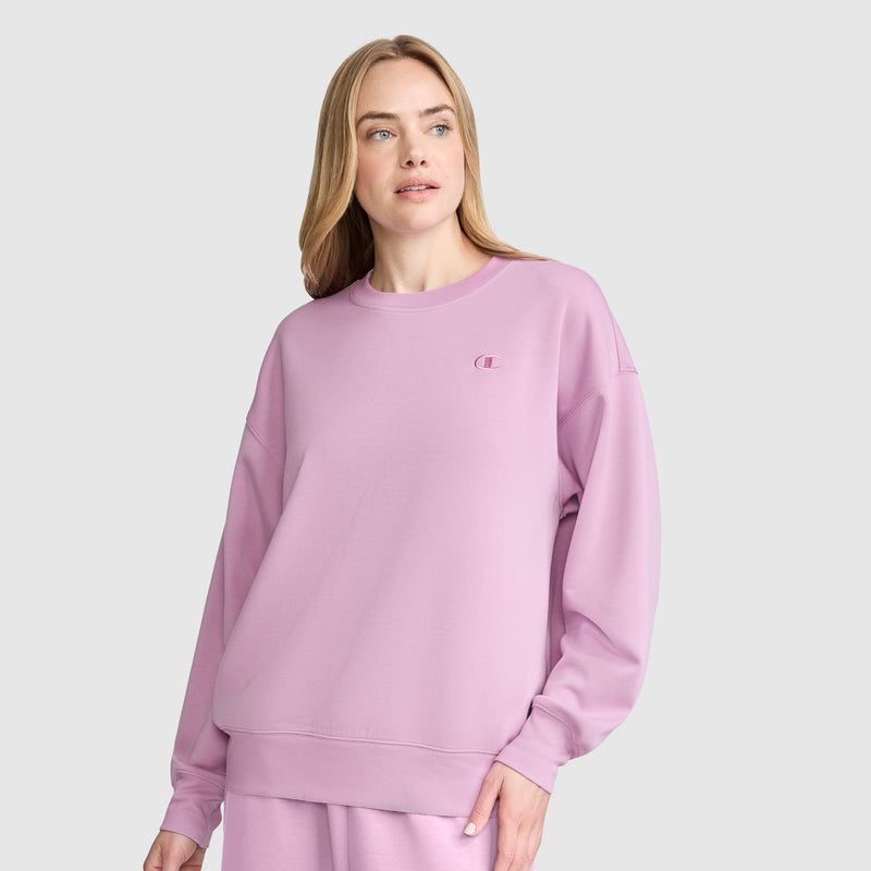 A woman stands against a plain light gray background, looking slightly to the side with a neutral expression, dressed in a loose-fitting pastel pink Champion Crewneck Pullover Sweatshirt and matching pants. The athletic silhouette of her Champion top adds a modern touch to her laid-back look.