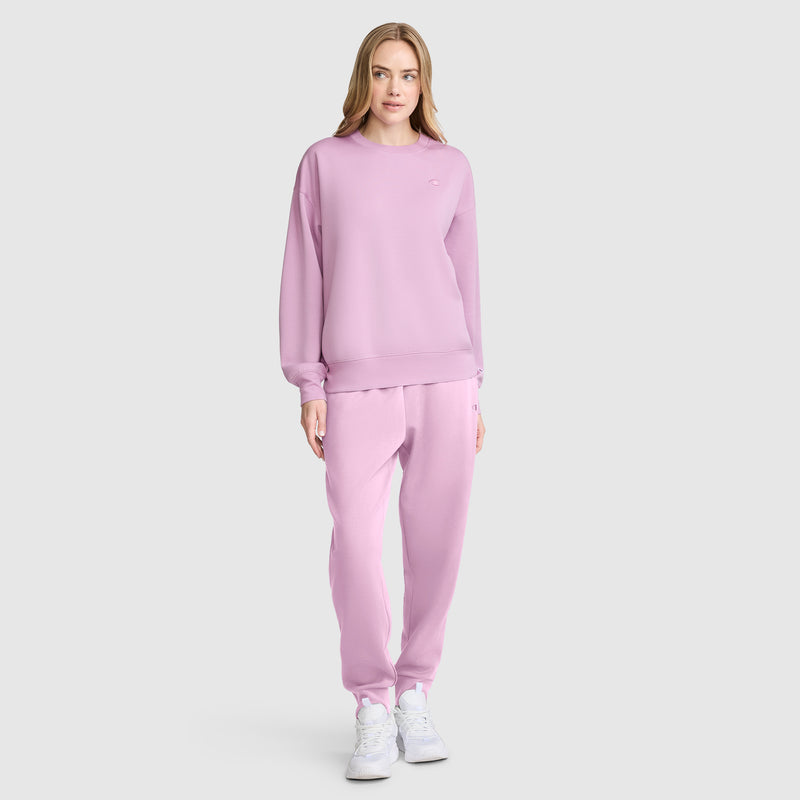 A woman stands facing forward against a plain light gray background, wearing a matching light pink Champion Crewneck Pullover Sweatshirt and sweatpants with white sneakers.