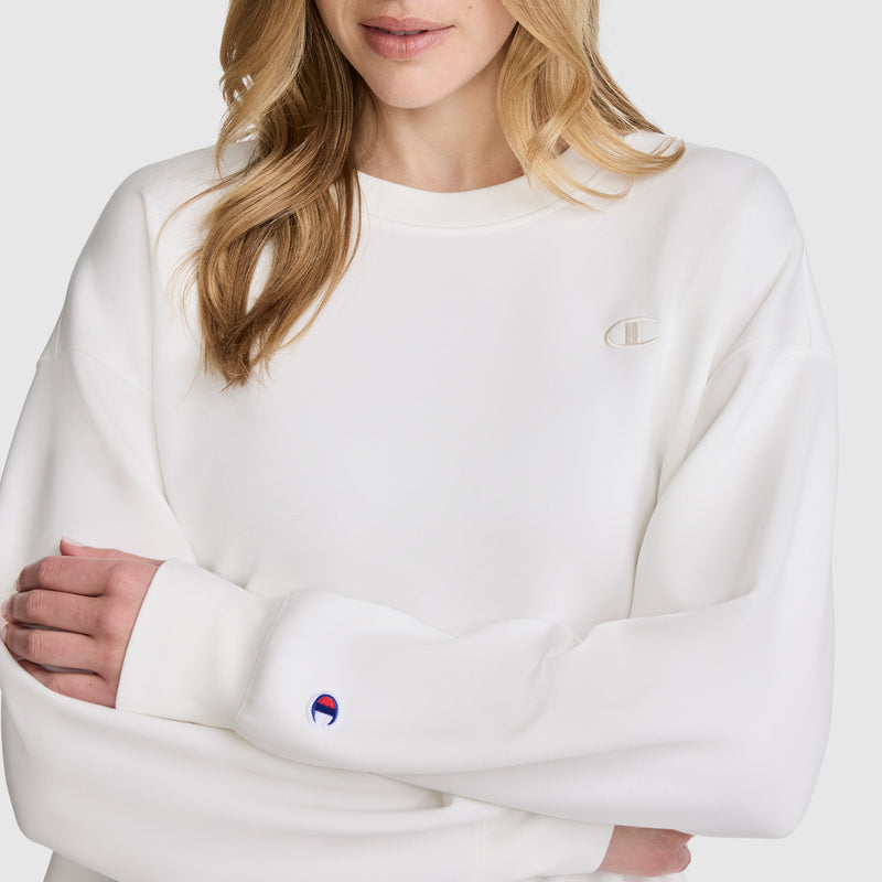 A woman with blonde hair, her arms crossed and only the lower half of her face visible, wears the Champion Crewneck Pullover Sweatshirt in white. Small Champion logos appear on the chest and sleeve, emphasizing its athletic silhouette.