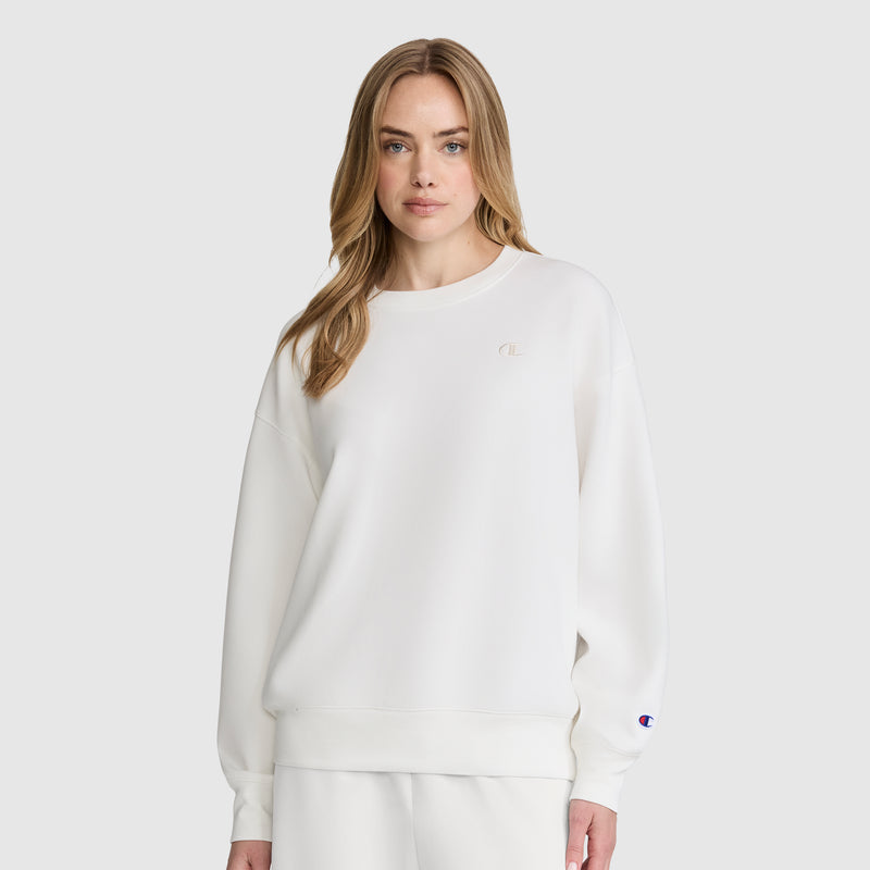A woman with long blonde hair wears the Champion Crewneck Pullover Sweatshirt in white, paired with matching white pants, creating an athletic silhouette as she stands against a plain light gray background.