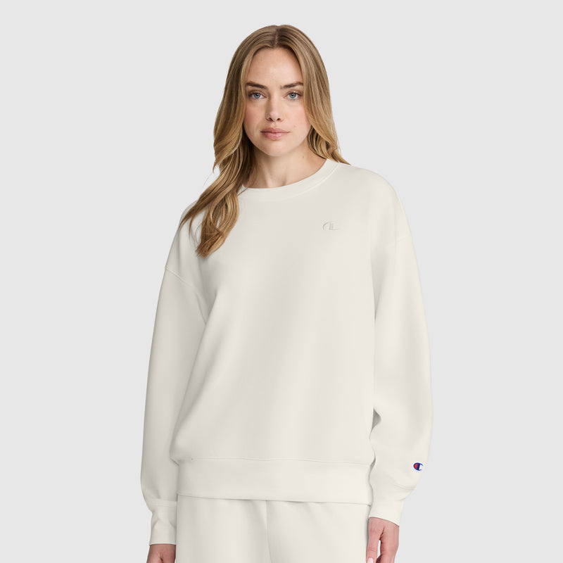 A woman wearing a cream-colored Champion Crewneck Pullover sweatshirt with a small logo on the chest and a patch on the sleeve.