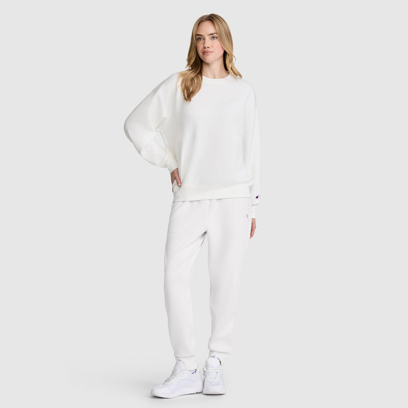 A woman stands against a plain light background, dressed in an athletic silhouette with a white modal scuba Crewneck Pullover Sweatshirt by Champion. She pairs it with matching sweatpants and white sneakers, her light brown hair complementing her relaxed pose as she effortlessly showcases the comfort and style of this Champion piece.