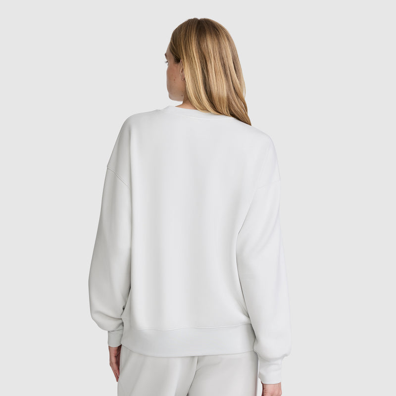 A woman with long, light brown hair is seen from behind against a plain, light background, wearing a loose-fitting Champion Crewneck Pullover Sweatshirt paired with matching white pants.