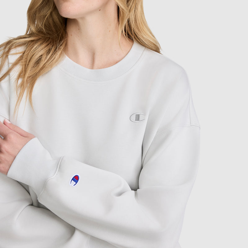 A woman wearing the Champion Crewneck Pullover Sweatshirt in light gray, featuring logo patches on the chest and sleeve, stands against a plain background with her arms crossed.
