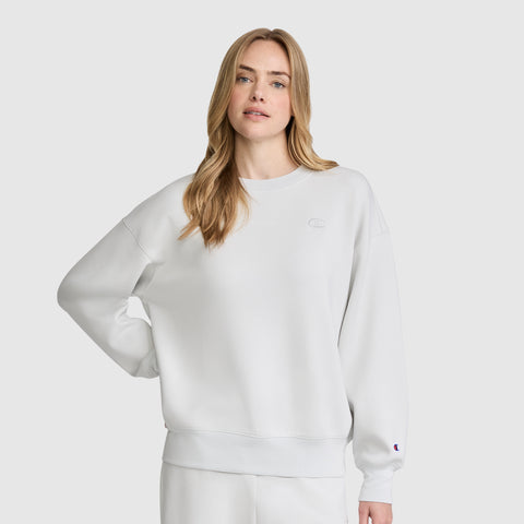 A woman with long blonde hair wears a loose-fitting white Champion Crewneck Pullover Sweatshirt and matching white pants, standing against a plain light gray background with one hand on her hip.