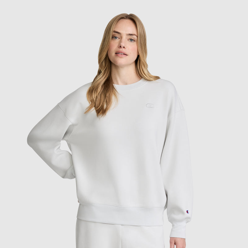 A woman with long blonde hair wears a loose-fitting white Champion Crewneck Pullover Sweatshirt and matching white pants, standing against a plain light gray background with one hand on her hip.
