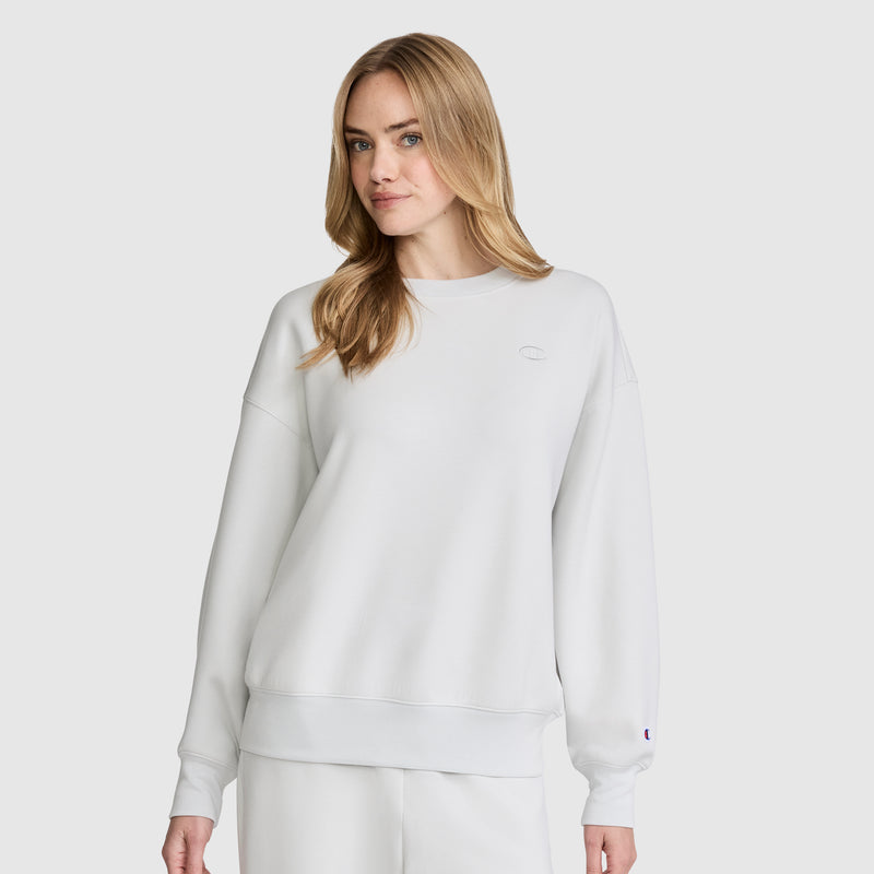 A woman with long blonde hair wears a plain white Champion Crewneck Pullover Sweatshirt and matching white pants, standing against a light gray background.