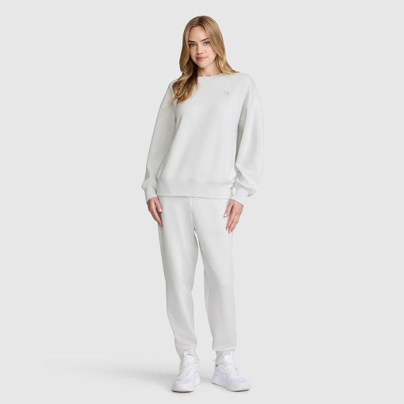 A woman stands against a plain white background wearing a light gray Champion Crewneck Pullover Sweatshirt, matching jogger pants, and white sneakers. She has blonde hair and a neutral expression, with one hand by her side and the other resting on her thigh.